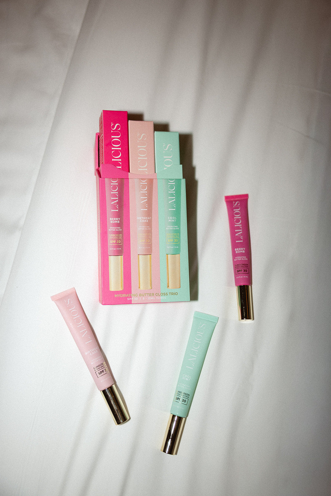 Three LALICIOUS lip balm tubes in Berry Bomb, Birthday Cake, and Cool Mint are arranged on a white fabric background next to their matching colored packaging box.