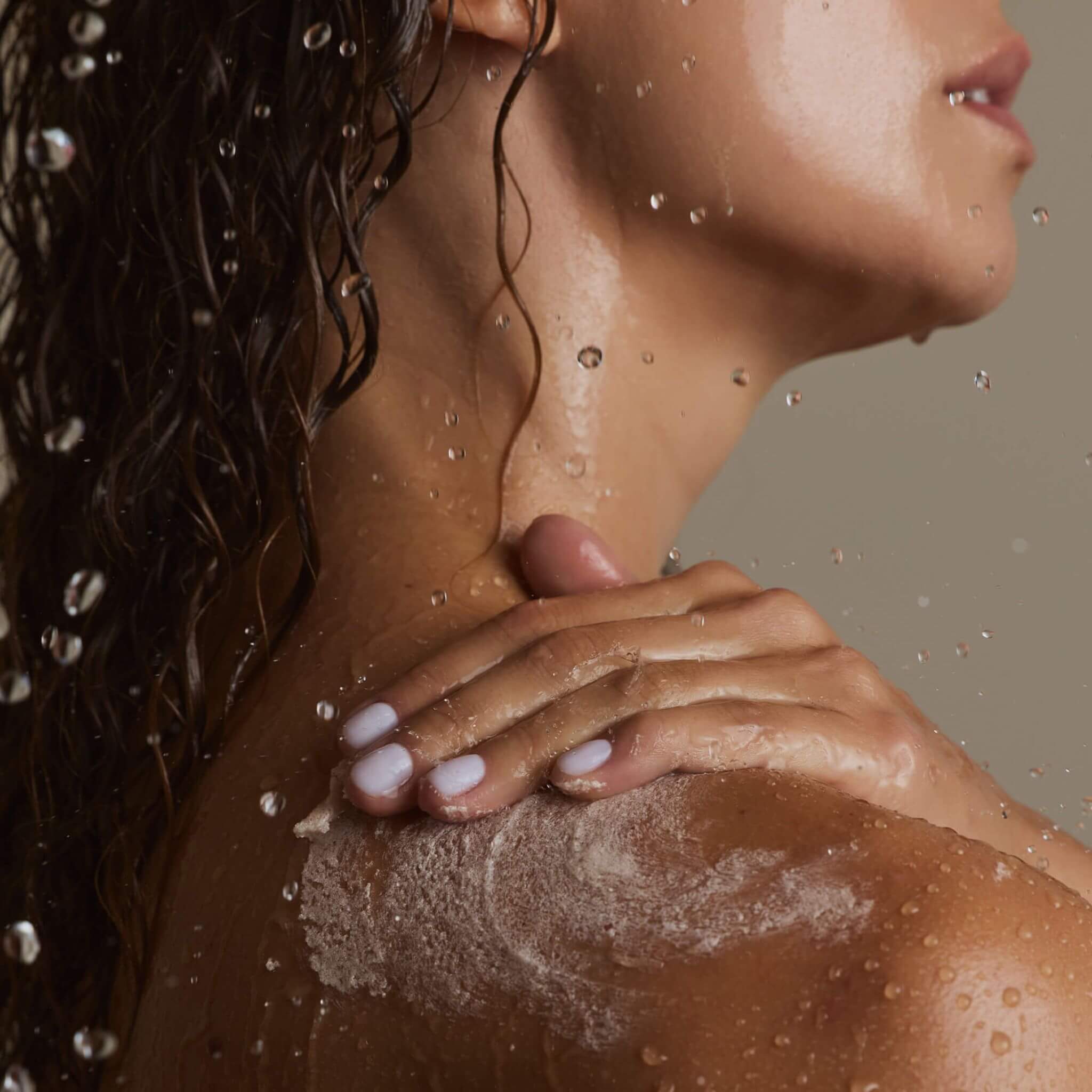 Close-up of a person with water droplets on their skin and hair, with a neutral background. Sugar scrub on shoulder of person.